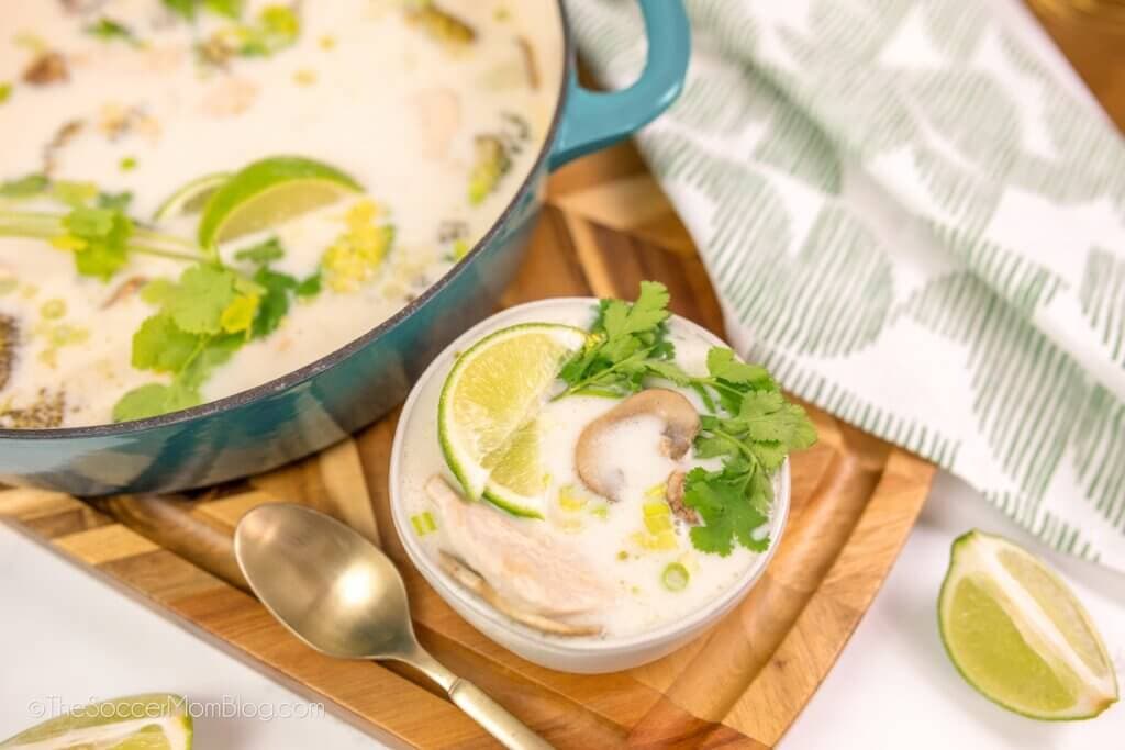 A bowl of creamy soup garnished with lime slices, cilantro, and mushrooms sits next to a pot of the same soup on a wooden board. A spoon and a lime wedge are nearby, with a green-and-white patterned cloth.