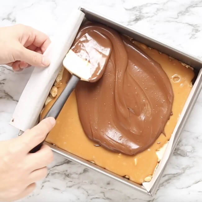 spreading melted chocolate on top of homemade snickers bar in baking pan