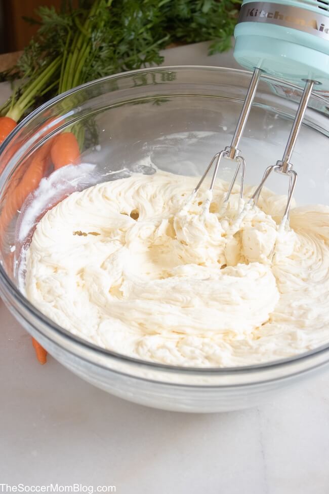 whipped white chocolate frosting in a glass mixing bowl, with electric mixer