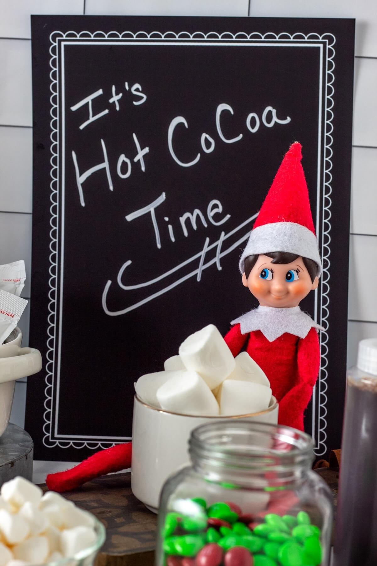 elf with a cup of hot cocoa and a chalkboard that says "It's hot cocoa time."
