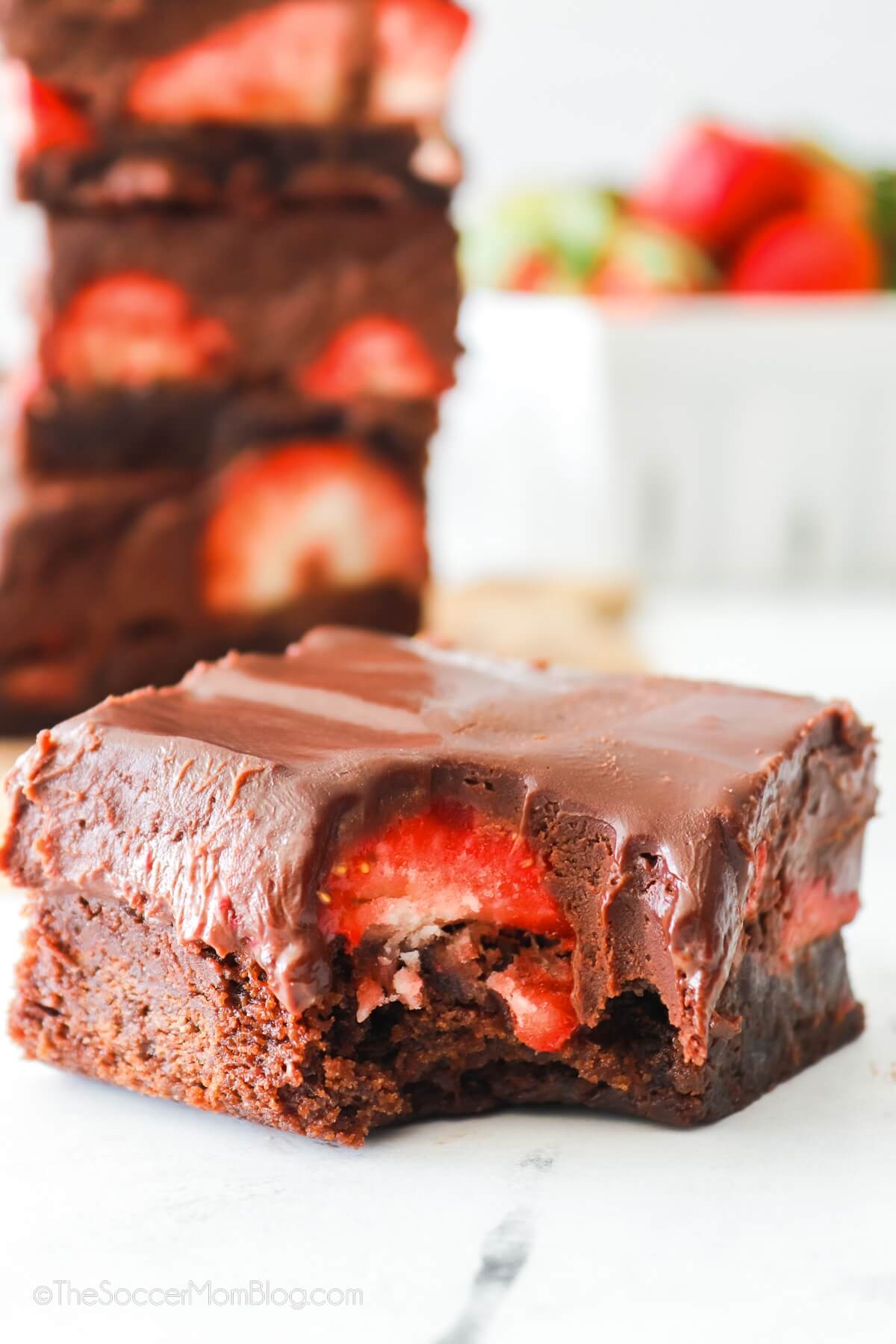 brownies topped with fresh strawberries and chocolate ganache, one with bite taken.