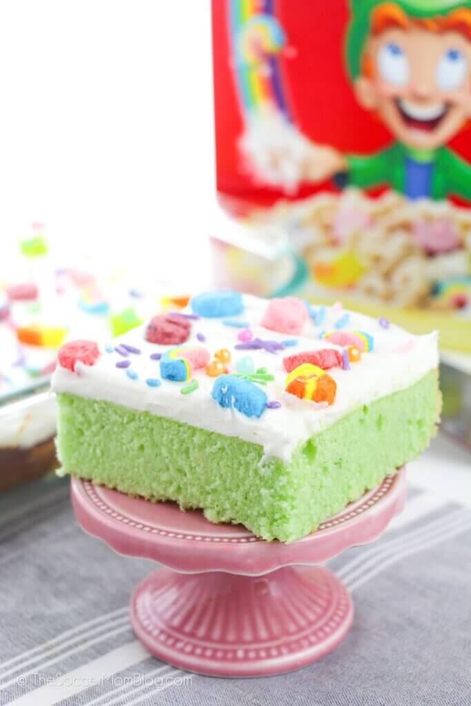 slice of Lucky Charms Cake on a pedestal, cereal box in background