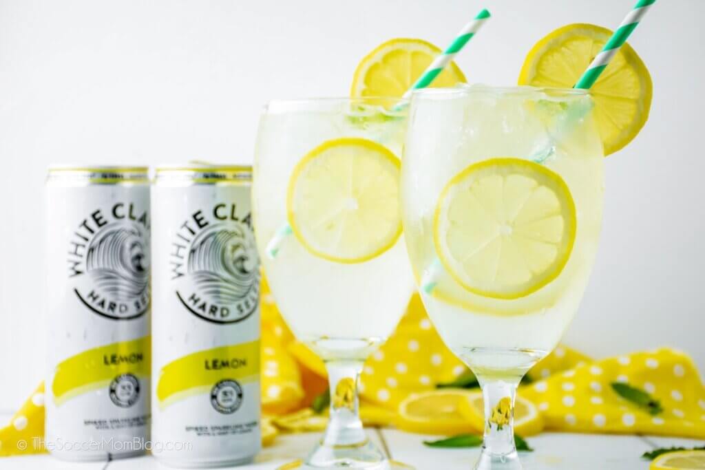 Two glasses of White Claw Sparkling Lemonade garnished with lemon slices, 2 cans of White Claw