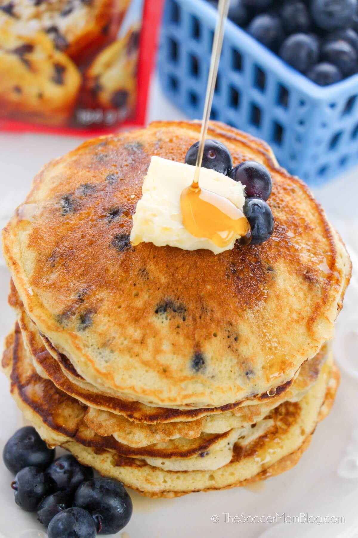 pouring syrup on blueberry pancakes, with crate of blueberries