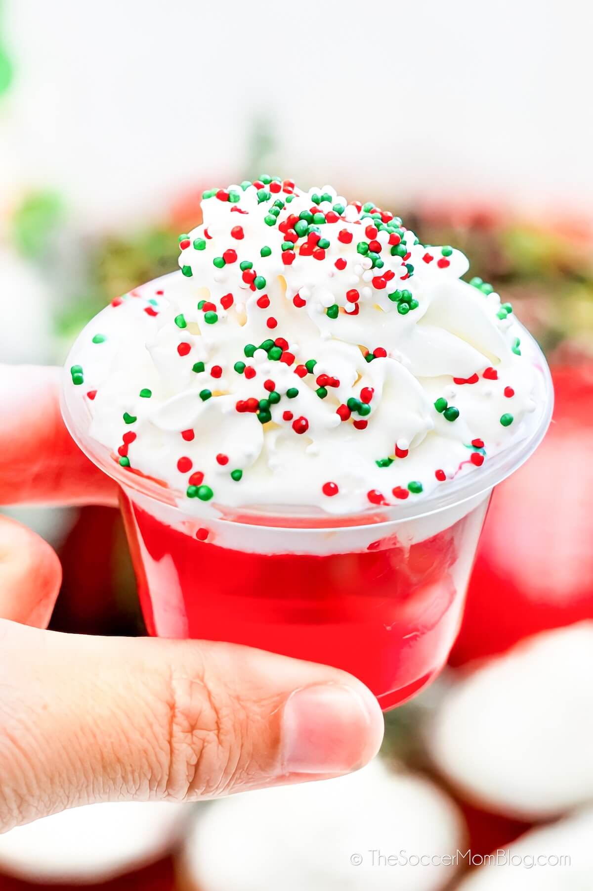 holding a red Christmas jello shot, with whipped cream and sprinkles