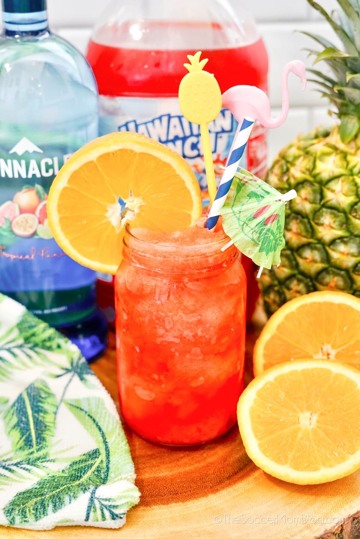 frozen Hawaiian punch cocktail in mason har, with vodka bottle, fruit punch jug, and fresh oranges