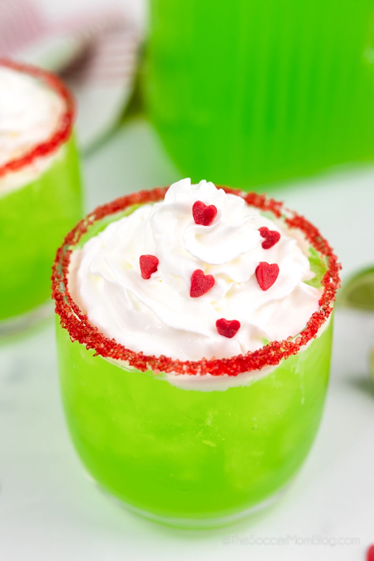 close up of a glass of green punch with whipped cream on top and red heart sprinkles.