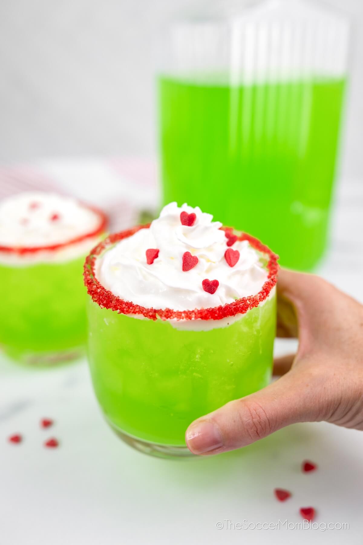 hand picking up a cup of Grinch punch that is bright green with whipped cream and red heart sprinkles.