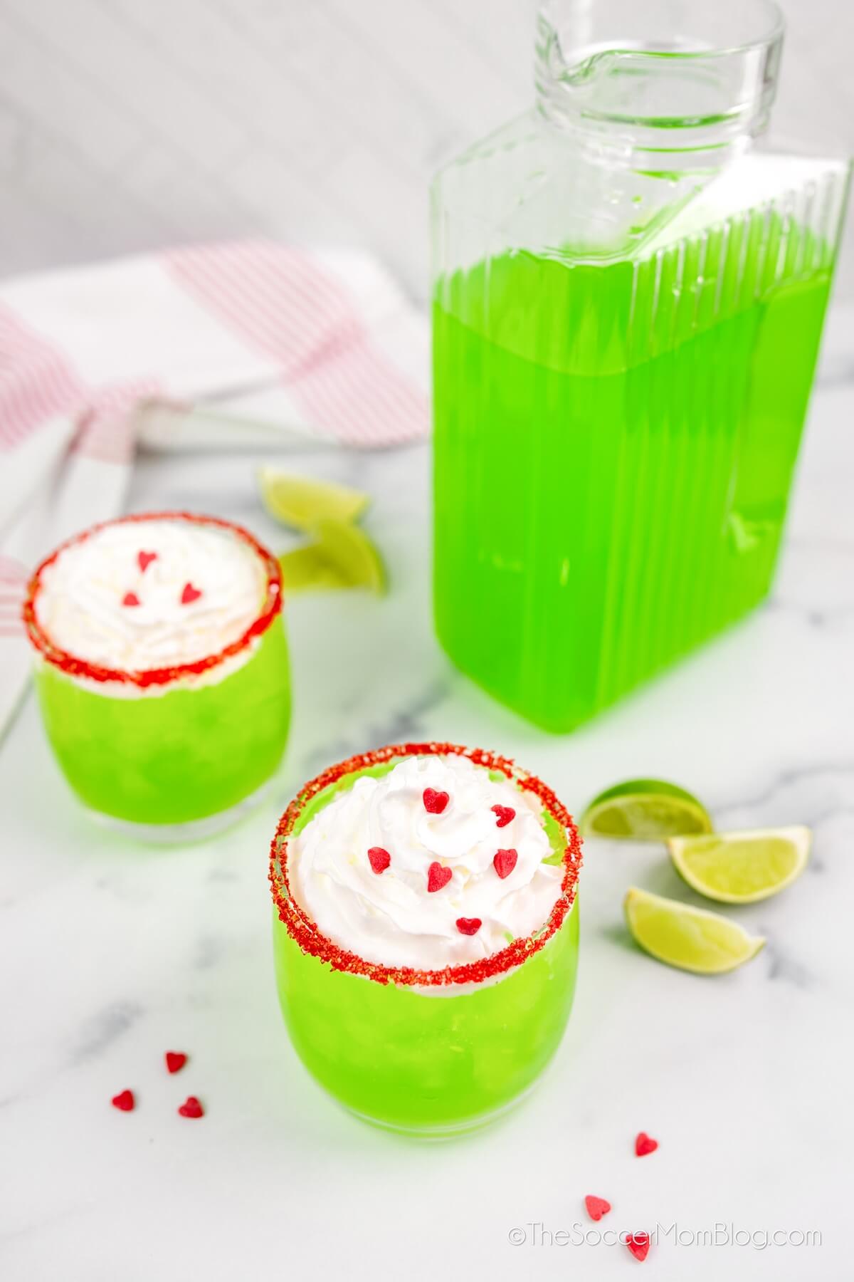 a full pitcher of lime green colored holiday punch, along with two individual glasses with whipped cream and sprinkles.