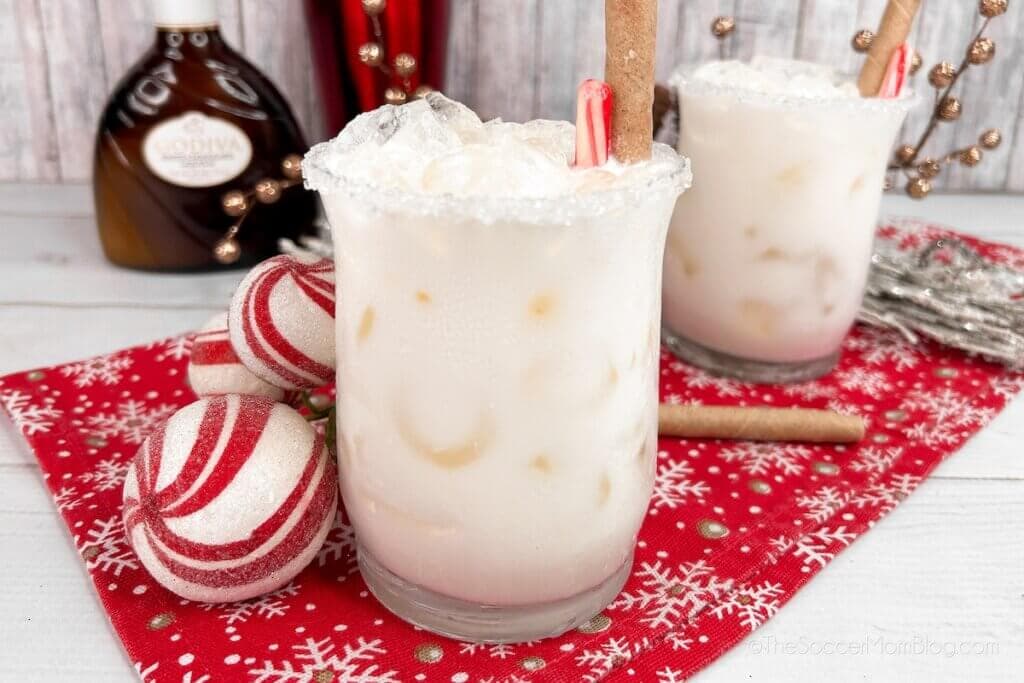Two glasses of creamy cocktail with ice and cinnamon sticks, on a red snowflake-patterned cloth. Candy canes garnish the rim. Holiday decorations and a bottle of liqueur are in the background.
