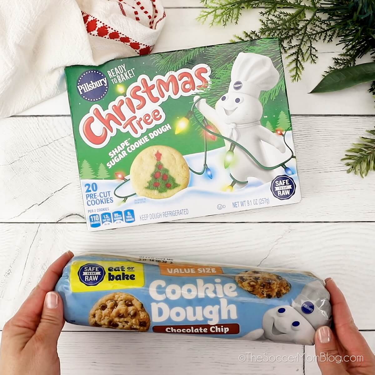 holding a roll of chocolate chip cookie dough and a box of Christmas sugar cookie dough on wooden workspace.