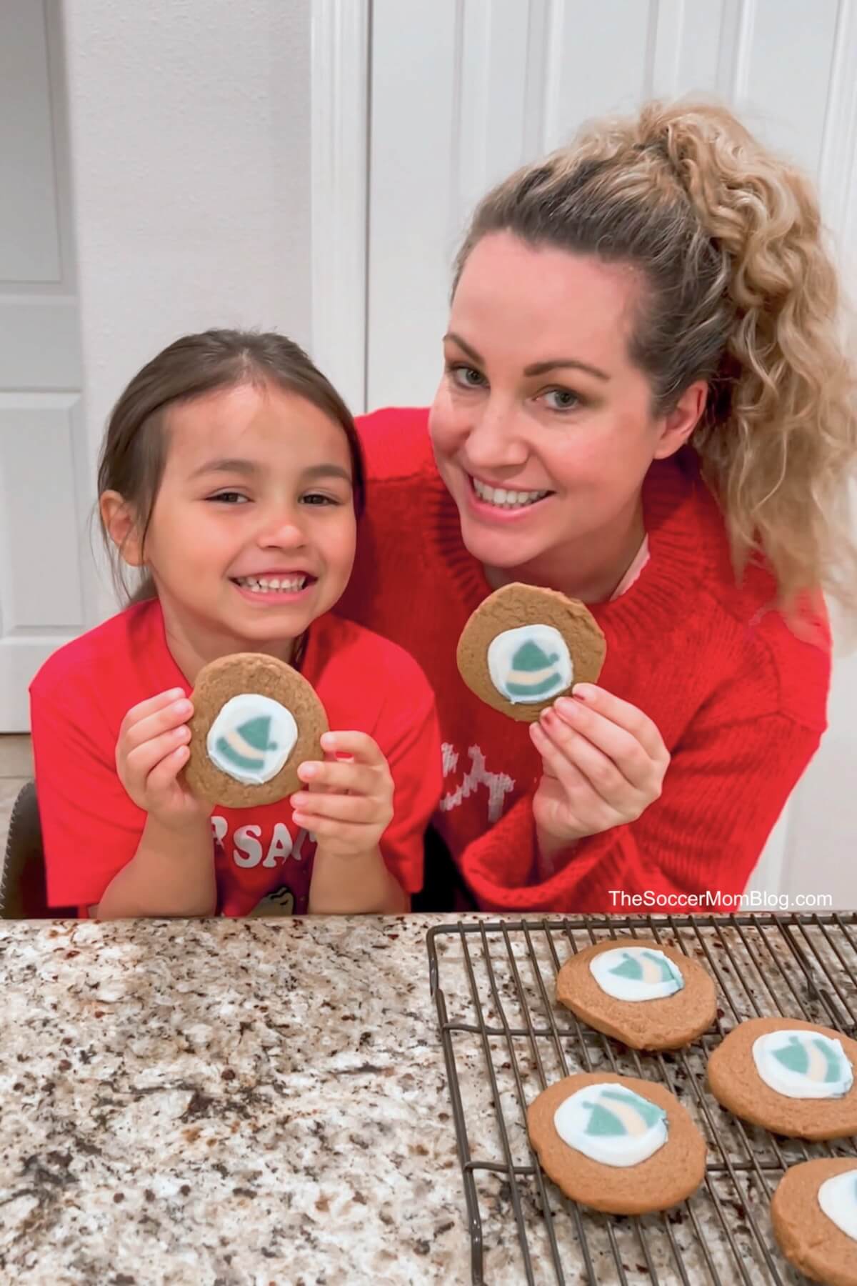 mom and daughter holding up gingerbread cookies with elf sugar cookies in the middle.