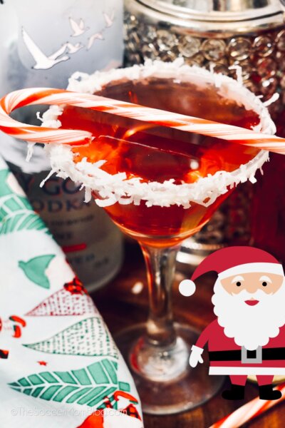 A red cocktail in a martini glass, with a coconut flake rim that looks like snow and a candy cane on top.