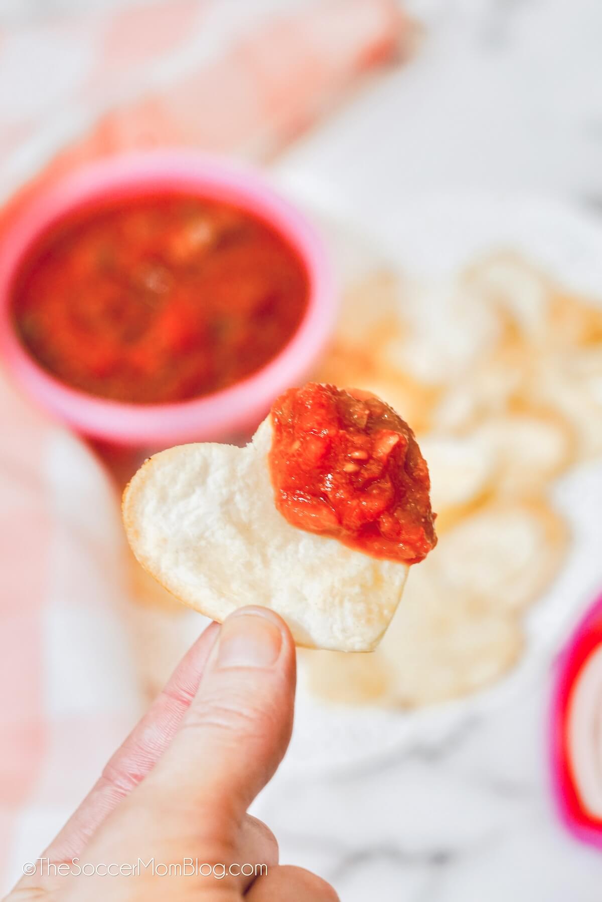 holding up a heart shaped tortilla chip, dipped in salsa.