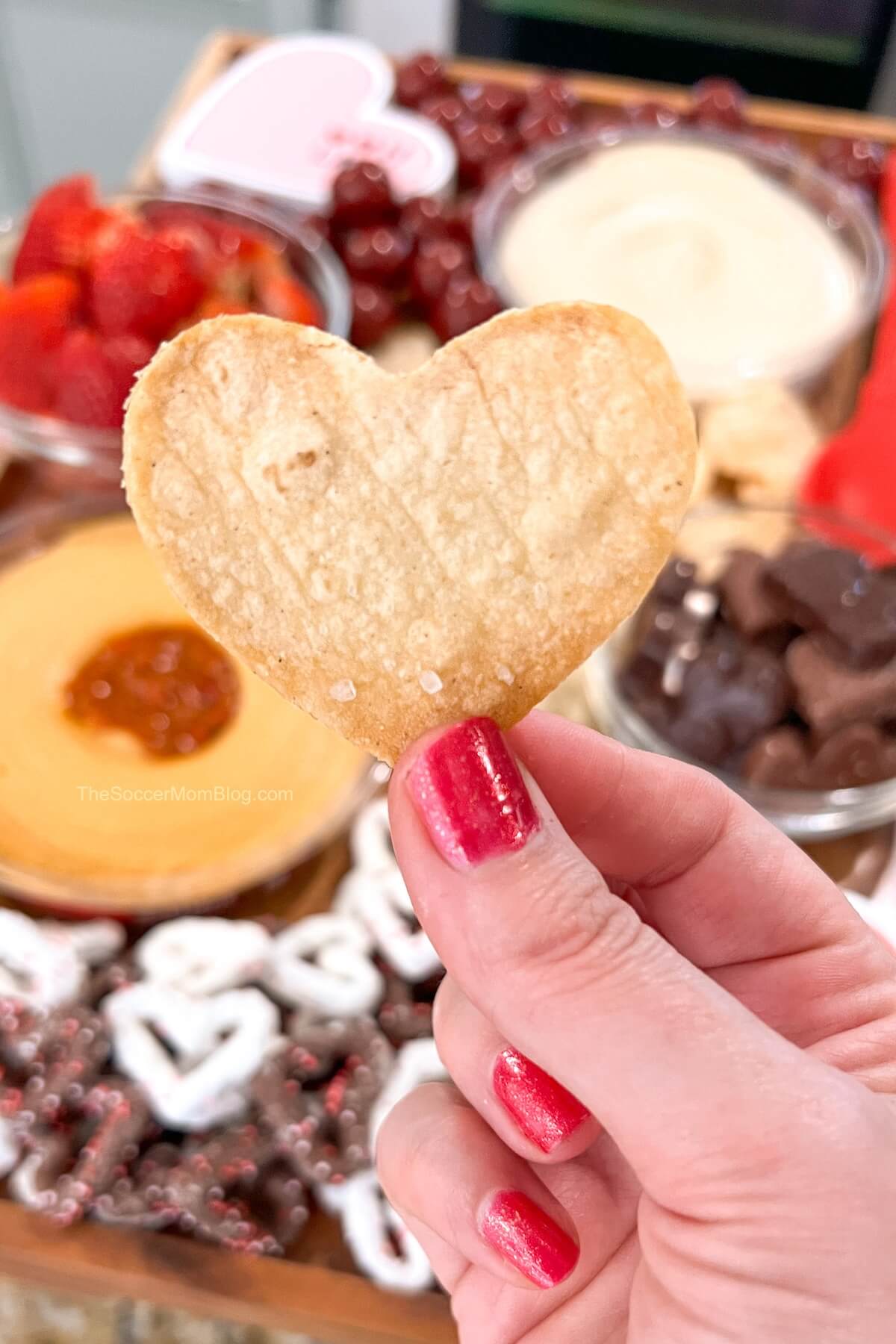 holding up a heart shaped tortilla chip in front of a Valentine snack board.