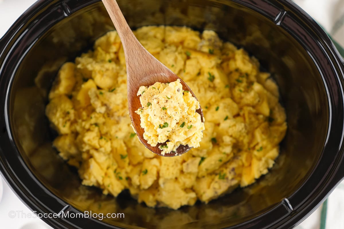 A wooden spoon lifting scrambled eggs out of a slow cooker.